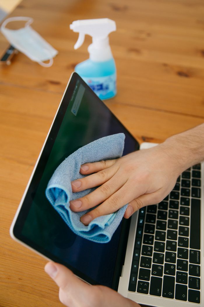 who-we-are A person wipes a laptop screen using a blue cloth and spray bottle on a wooden table for hygiene.